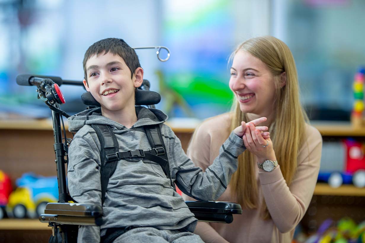 Photo of a boy in a power wheelchair with his mother smiling and holding his hand next to him.